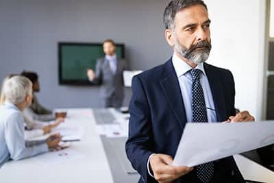 Businessman in blue suit stands with back to a room full of employees meeting